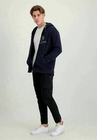 Ung mand iført en marineblå zip-up hættetrøje, hvid t-shirt, sorte cargo-bukser og hvide sneakers, stående med hænderne i lommerne.