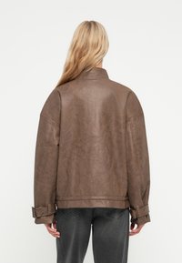Brązowa kurtka z sztucznej skóry z wysokim kołnierzem, luźnym krojem i detalami na mankietach. Posiada gładką teksturę i minimalistyczny design.