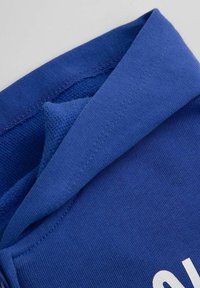 Niebieska tkanina z prążkowanym kołnierzem i gładką teksturą, z białym wzorem na powierzchni. Widoczne przeszycia na krawędziach.