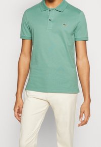 Polo vert en coton, avec un col, des manches courtes et un petit logo brodé. Porté avec un pantalon beige clair.