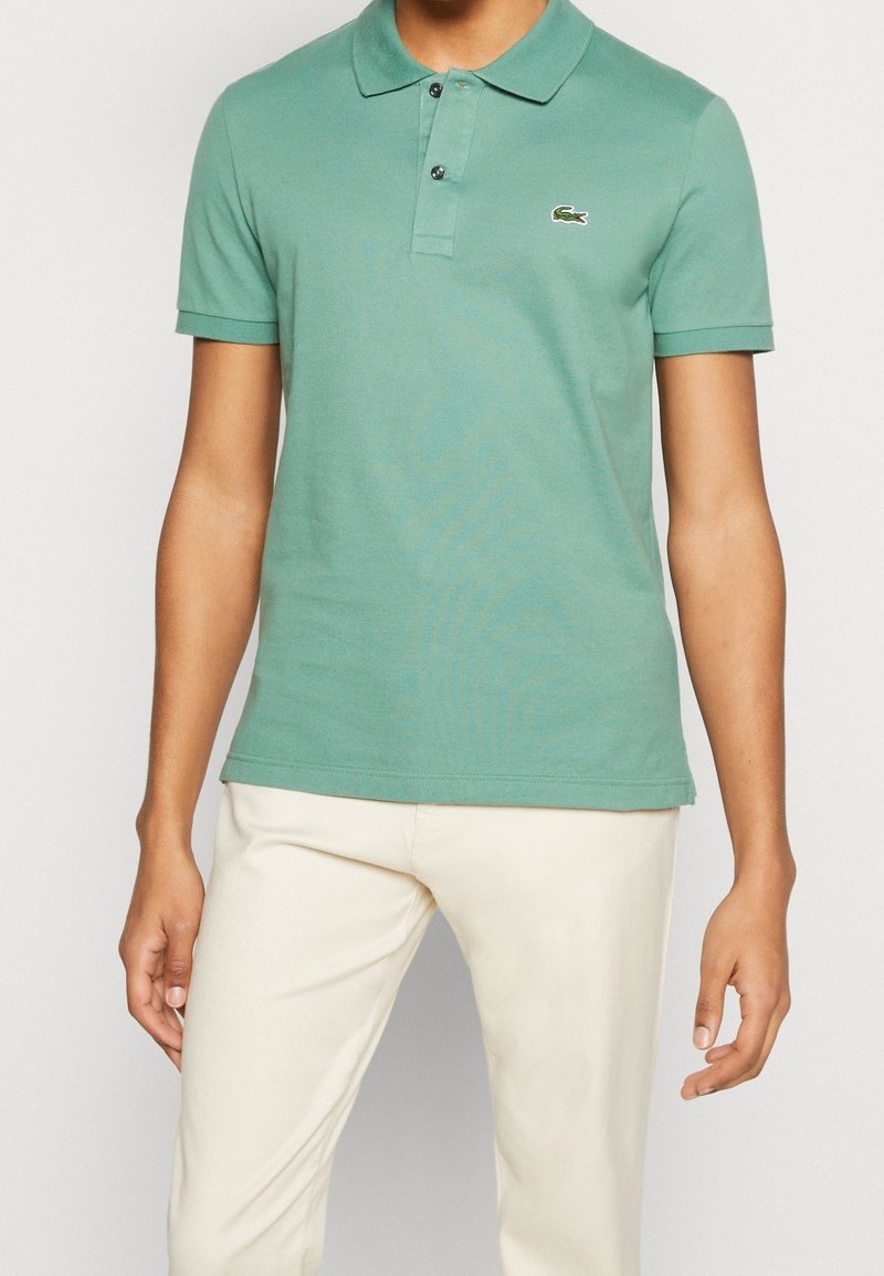 Polo vert en coton, avec un col, des manches courtes et un petit logo brodé. Porté avec un pantalon beige clair.
