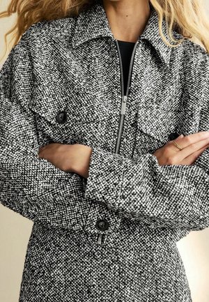 Kobieta w czarno-białej, teksturowanej kurtce na zamek z kołnierzem i kieszeniami z patkami zapinanymi na guziki, ze skrzyżowanymi ramionami, pokazująca złoty pierścionek na lewej dłoni.