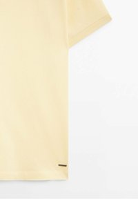 Chemise jaune clair à manches courtes avec une petite étiquette rectangulaire métallique près de l'ourlet inférieur sur fond blanc.