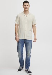 Kortermet, lysebeige skjorte med camp-krage, knapplukking foran, kombinert med blå jeans og svarte sneakers. Enkel design og stoff.