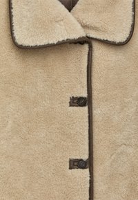 Manteau en shearling beige avec une texture douce, bordure brun foncé et deux boutons de fermeture. Doté d'un large col et de détails cousus sur toute sa surface.
