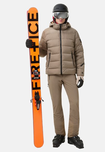 Polstrovaná béžová lyžařská bunda s kapucí, doplněná o lyžařské kalhoty barvy ovesné kaše. Drží oranžovou lyži s černým písmem. Součástí jsou černé rukavice a helma.
