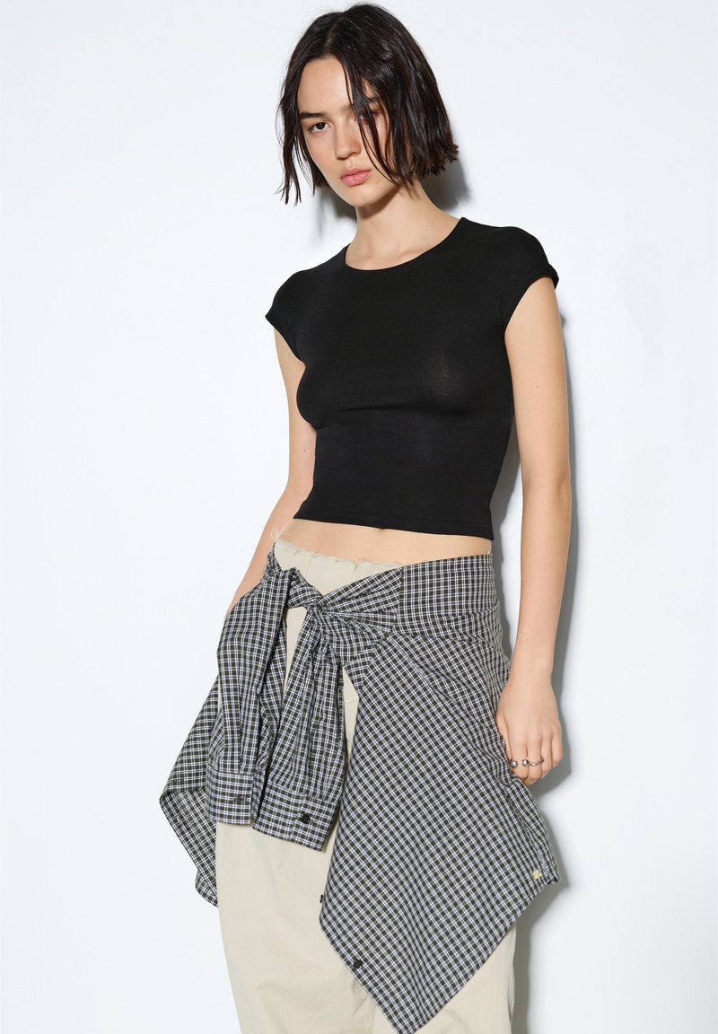 Schwarzes eng anliegendes Crop-Top mit kurzen Ärmeln, kombiniert mit einem karierten Wickelrock mit schwarz-weißen Karomustern. Beige hoch taillierte Hose.