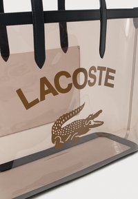 Prozirna torba od plastike s smešnim 'LACOSTE' logom i grafikom krokodila, crnim kožnim obrubima i ručkama, pravokutnog oblika, glatke teksture.
