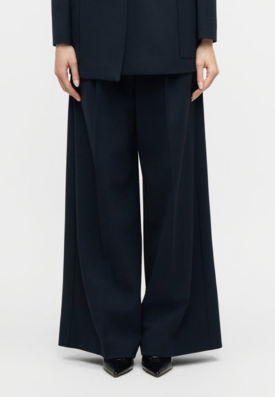 Personne portant un pantalon large noir et un blazer assorti, debout les mains détendues le long du corps, chaussures noires pointues visibles.