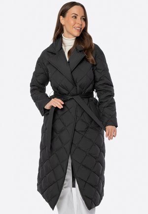 Vrouw draagt een lange zwarte doorgestikte jas met ceintuur over een witte coltrui en witte broek, glimlacht en stelt de jasceintuur bij.