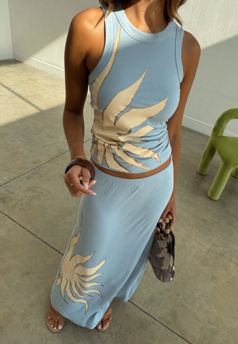 Femme portant un débardeur bleu clair et une jupe avec des motifs de soleil beiges, tenant un sac à motifs avec des perles en bois dans un cadre intérieur minimaliste.