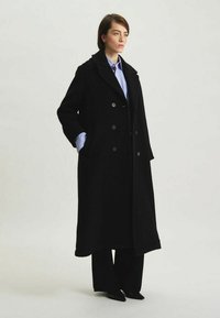 Manteau noir croisé en tissu texturé, avec des revers crantés, des poches latérales et une silhouette longue. Associé à une chemise bleue et un pantalon.