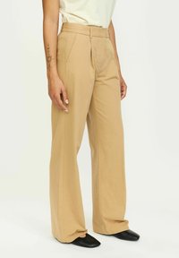 Bež hlače širokih nogavica s visokim strukom, koje imaju prednje nabore, glatku teksturu i blago flared nogavice, u kombinaciji s crnim cipelama na špic.