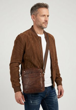 Mand iført brun ruskindjakke, hvid skjorte, blå jeans og bærende en brun læder crossbody taske med mærket "VENEZIA" mod en neutral baggrund.