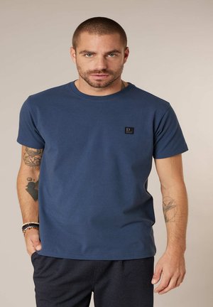 Man met kort haar en baard draagt een blauw T-shirt met logopatch, donker patroonbroek, armbanden aan de linkerpols en heeft zichtbare tatoeages op de arm.