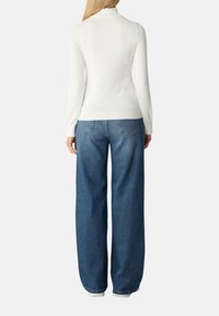 Femme portant un haut à manches longues blanc et un jean bleu ample, se tenant de dos sur un fond blanc uni.