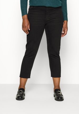 Persoon die een korte zwarte spijkerbroek draagt, zwarte loafers met zilveren gespen en een donker groene trui met lange mouwen, staand tegen een witte achtergrond.