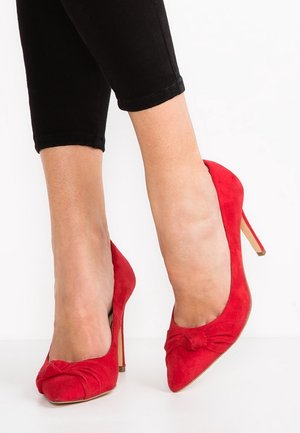 Czerwone zamszowe szpilki z wysokim obcasem, zakończone spiczastym noskiem z detalem w postaci kokardki na przodzie. Obcas typu szpilka i fakturowana powierzchnia. Noszone z czarnymi obciętymi spodniami.
