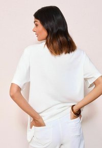 Haut blanc à manches courtes en tissu doux, avec une coupe décontractée. Les cheveux du modèle tombent légèrement au-delà des épaules, coiffés avec un effet ombré.