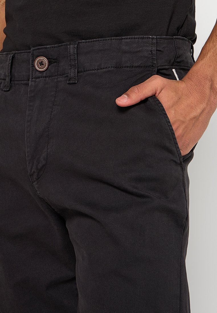 Schwarze Baumwollhose mit geradem Schnitt, ausgestattet mit einer Vordertasche und einem braunen Knopfakzent am Bund. Dezente Texturdetails sichtbar.