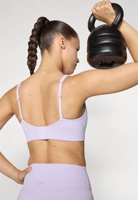 Lilla idretts-BH med justerbare stropper og hekklukking, som fremhever et glatt, stretchy stoff. En kvinne løfter en svart kettlebell.