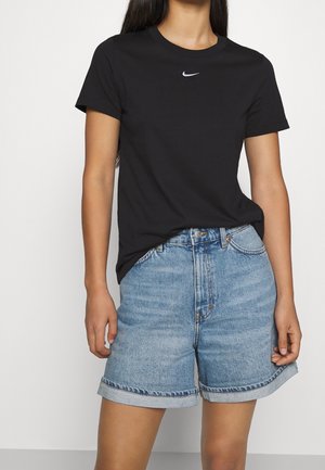 Kobieta ubrana w czarną koszulkę Nike i jasnoniebieskie szorty jeansowe z wysokim stanem na jednolitym białym tle.