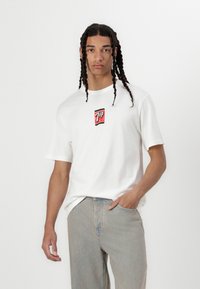 Hvid bomulds t-shirt med et 7UP-logo i rød og sort i midten, parret med lyseblå jeans og stylet med fletninger.