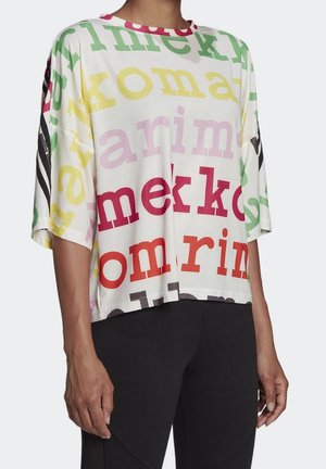 Loses weißes T-Shirt mit übergroßen, mehrfarbigen Buchstaben und halblangen Ärmeln, getragen mit eng anliegenden schwarzen Hosen an einer stehenden Person.