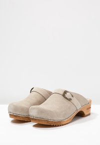Beige Wildlederschuhe mit einer Holzsohle, runder Zehenform und einem Band über der Oberseite, das mit einer Metallbuckle verziert ist. Strukturierte Oberfläche.
