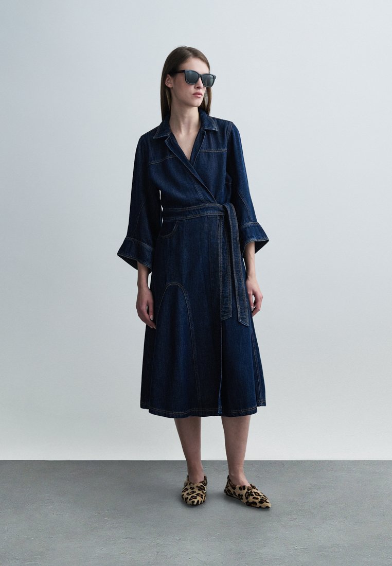 Robe portefeuille en jean avec manches trois-quarts, taille cintrée et jupe évasée. Portée avec des ballerines à motif léopard et des lunettes de soleil foncées.