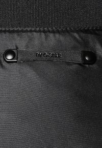 Svart tyg med en slät struktur har en sydd etikett märkt "BY G-STAR." Den har även svart tryckknapp för säker stängning.