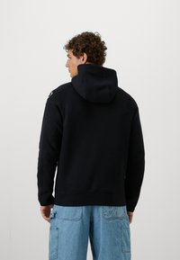 Sort hoodie med en stor hætte, ribbede manchetter og kant, der har et diskret logo på skulderen. Modellen har lyseblå cargo jeans på.