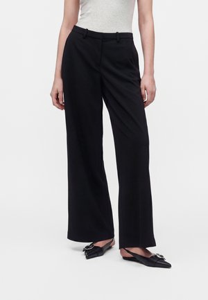 Femme portant un pantalon large noir taille haute et des ballerines noires à bride arrière pointues avec un détail de boucle argentée sur fond blanc.