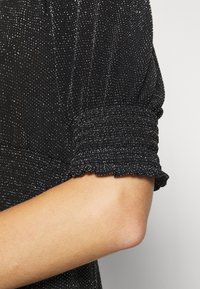 Svart topp i en strukturerad tygkvalitet med silverglittrande detaljer. Korta, volangärmar med samlade manschetter, närbild av armen och ärmen.