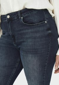 Jeans de mezclilla oscuros de corte alto y ajustado. Presentan bolsillos frontales, un pequeño detalle desgastado y herrajes metálicos plateados en la cintura.