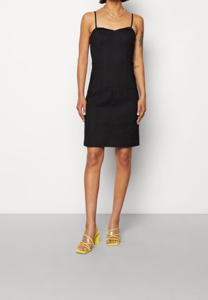 Robe noire sans manches avec une silhouette ajustée, mettant en valeur un décolleté en cœur et des coutures verticales. Associée à des talons jaunes à lanières.