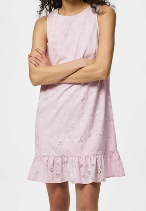Femme portant une robe sans manches rose clair avec un motif floral ajouré et un ourlet à volants, les bras croisés sur la poitrine.