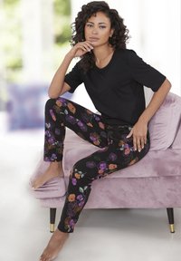 Top negro de manga corta combinado con pantalones negros con patrones florales coloridos en púrpura, naranja y verde, sentado en una silla de terciopelo rosa.