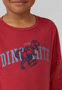 Rotes Langarm-T-Shirt mit einem Dinosaurier-Graphic in Blau und Schwarz, mit dem Wort "DINO-MITE", das prominent darunter angezeigt wird.