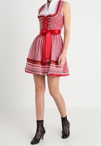 Červená a bílá vzorovaná dirndl šaty s přiléhavým korzetem, dekoračními knoflíky a širokým červeným saténovým opaskem. Černé kotníkové ponožky a podpatky.
