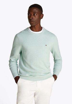 Man draagt een lichtblauwgroene, getextureerde trui met lange mouwen en een witte broek, handen in de zakken, kijkt opzij tegen een witte achtergrond.