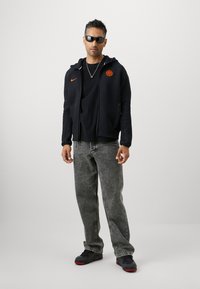 Sudadera negra con cremallera y logo de Nike naranja, camiseta negra, vaqueros de mezclilla grises y zapatillas negras con detalles rojos. Gafas de sol y detalles de joyería.