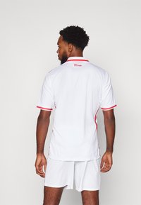 Jersey atlético blanco con detalles rojos, mangas cortas y un cuello. La parte trasera presenta un logo cerca del cuello, acompañado de pantalones cortos blancos.