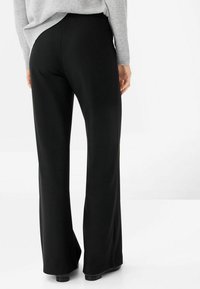 Kvinna i svarta vida byxor och en ljusgrå tröja, stående med ryggen mot kameran framför en enfärgad vit bakgrund.