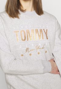 Sudadera gris claro con escote redondo, que presenta gráficos metálico dorados "TOMMY JEANS" y estrellas. Fabricada con una suave tela texturizada.