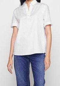 Kvinna som bär en lös vit blus med korta ärmar och krage, kombinerad med blå jeans, stående mot en enkel ljus bakgrund.