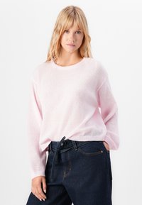 Woman with long blonde hair wearing a pale pink knit sweater tucked into high-waisted dark blue jeans with a tied belt.