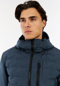 Jonge man met donker haar die een dichtgeritste marineblauwe high-performance pufferjack met capuchon draagt tegen een effen witte achtergrond.