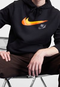Svart hoodie i bomull med en stor Nike-logotyp i gradient orange och gult samt en mindre svart Nike-logotyp. Modellen sitter på en kromstol.