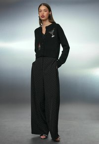 Cardigan negru tricotat cu motive de cocktail brodate, asociat cu pantaloni largi negri cu buline albe. Material texturat și nasturi.